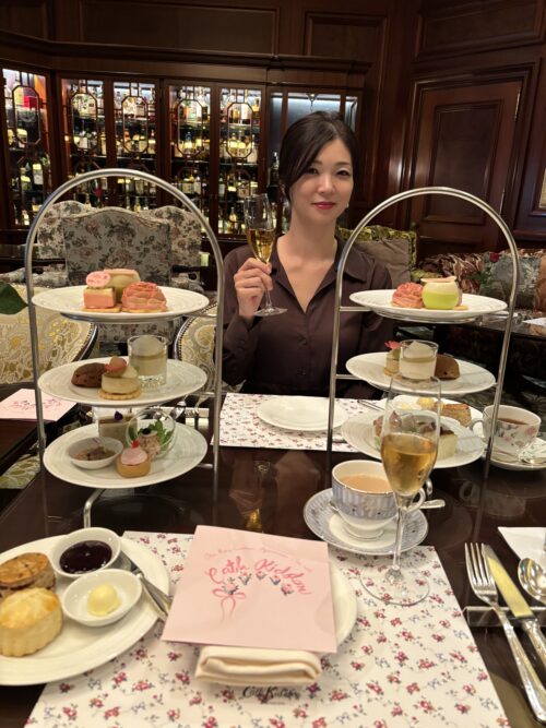 クラシカルなホテルのラウンジで、笑顔のアジア人女性がグラスシャンパンを持ち、手前には豪華な3段ティースタンドが並べられ、可愛らしいスイーツやセイボリー、スコーンが美しく盛り付けられている。テーブルには花柄のランチョンマットとピンクのメニューカードがある。