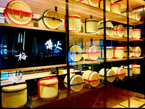 鉄板焼きレストラン「川梅 鉄板 TENKA（てんか）」の店内の装飾。日本酒の樽を模したオブジェが壁一面に飾られ、和モダンな雰囲気。中央のモニターに「川梅」「傳火 TEPEAN TENKA」のロゴが表示されている。