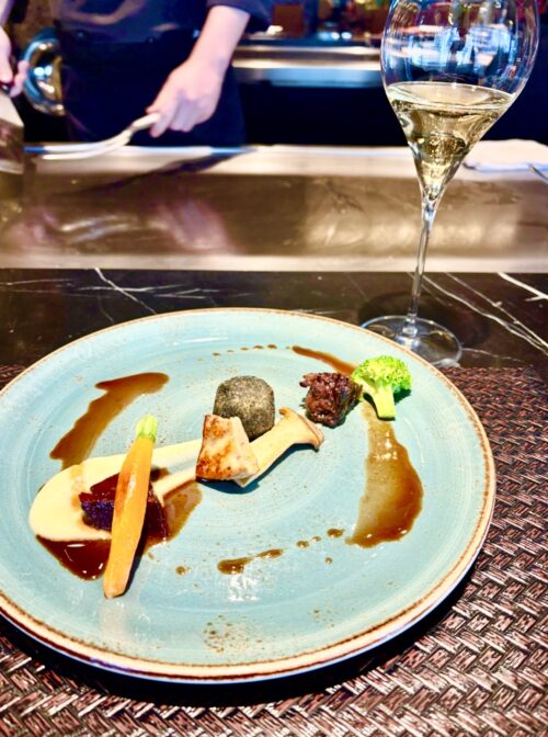 鉄板焼きカウンターで、シェフが調理する様子を背景に、青緑色の皿に美しく盛り付けられたタンシチュー、トリュフクロケット、野菜の前菜。手前にはシャンパンのグラスがある。