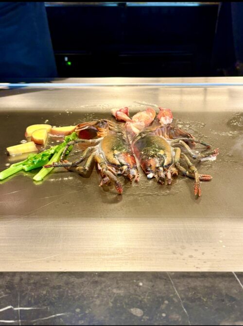 鉄板カウンターの上で、調理中のオマール海老。頭と殻が左右に広げられた状態で、エビの身、アスパラガスなどの野菜と共に焼かれている。