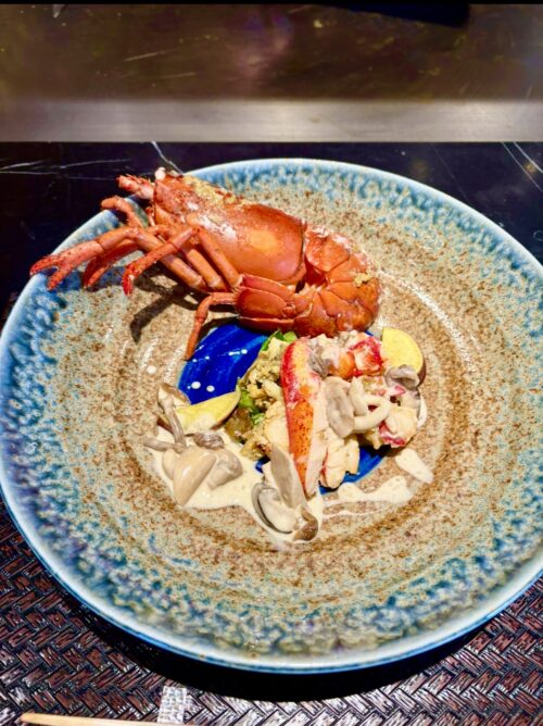 濃厚な青と茶の陶器皿に盛り付けられた、オマール海老のソテー。殻付きの海老と、海老の身、きのこ、クリームソースが美しく添えられている。