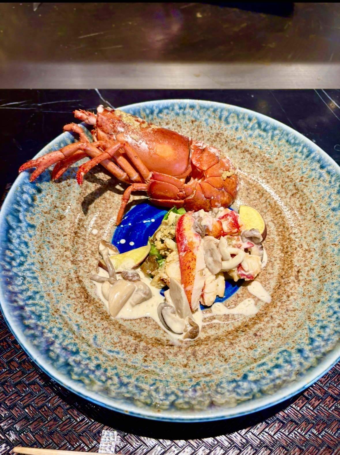 濃厚な青と茶の陶器皿に盛り付けられた、オマール海老のソテー。殻付きの海老と、海老の身、きのこ、クリームソースが美しく添えられている。