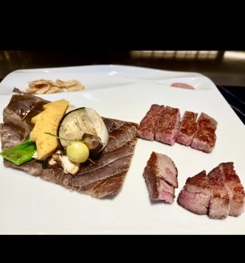 白い皿に盛り付けられた、火入れが完璧な和牛のステーキ（フィレまたはサーロイン）。付け合わせの焼き野菜は、大きな朴葉の上に配置されている。