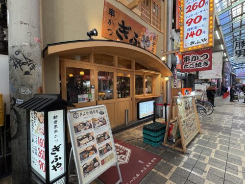 商店街にある、木造の和風建築の串かつ店「ぎふや」の正面外観。店頭には、生ビールや串かつの価格を示す縦長の看板と、朝食メニューの立て看板が置かれている。