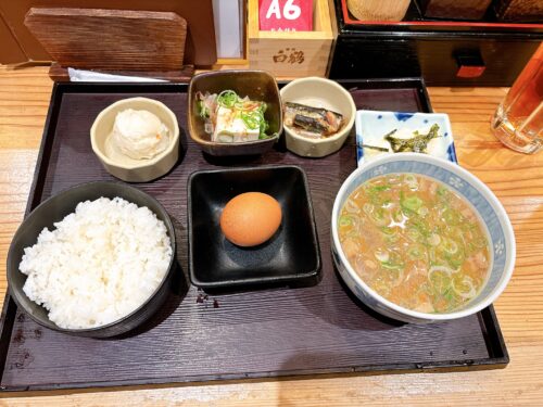 黒いお盆の上に並べられた和食の朝食セット。左から白ご飯、中央に生卵、右に豚汁の入った器が配置され、その他に豆腐、魚の煮物、ポテトサラダなどの小鉢が複数並んでいる。